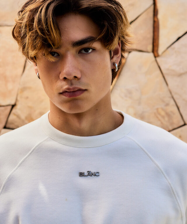 Close-up of a model wearing a white ribbed crewneck top, featuring a metallic BLÃNC logo badge on the chest.