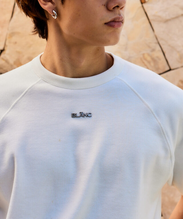 Close-up of a model wearing a white ribbed crewneck top, featuring a metallic BLÃNC logo badge on the chest.