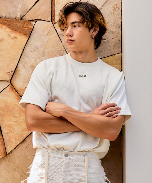 Close-up of a model wearing a white ribbed crewneck top, featuring a metallic BLÃNC logo badge on the chest.